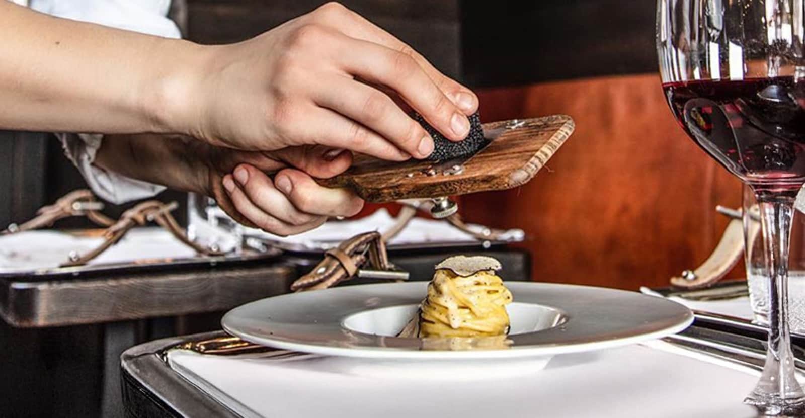 Mano che rifinisce con lamette di tartufo nero un piatto di pasta al centro di un grande piatto bianco.