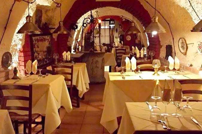 Sala da pranzo con soffitto a volta in pietra, tavoli con tovaglie bianche e lampade a cono pendenti.