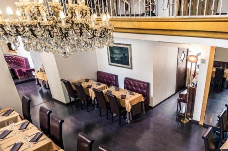 Sala vista dall'alto con lampadario di cristallo, tavoli con divanetti in pelle bordeaux e balconata al piano superiore.