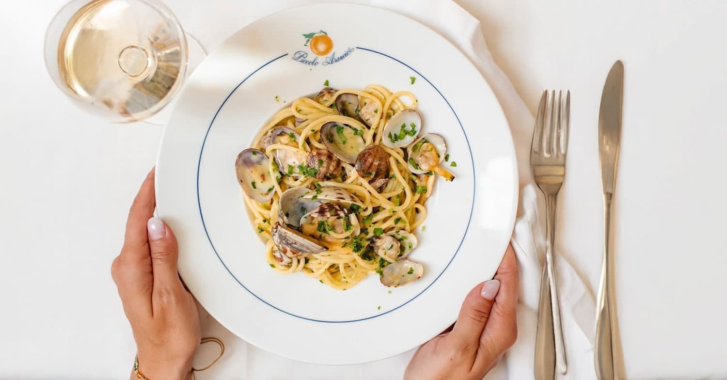 Piatto di spaghetti alle vongole tenuto con le mani, bicchiere di vino bianco e posate ai lati.