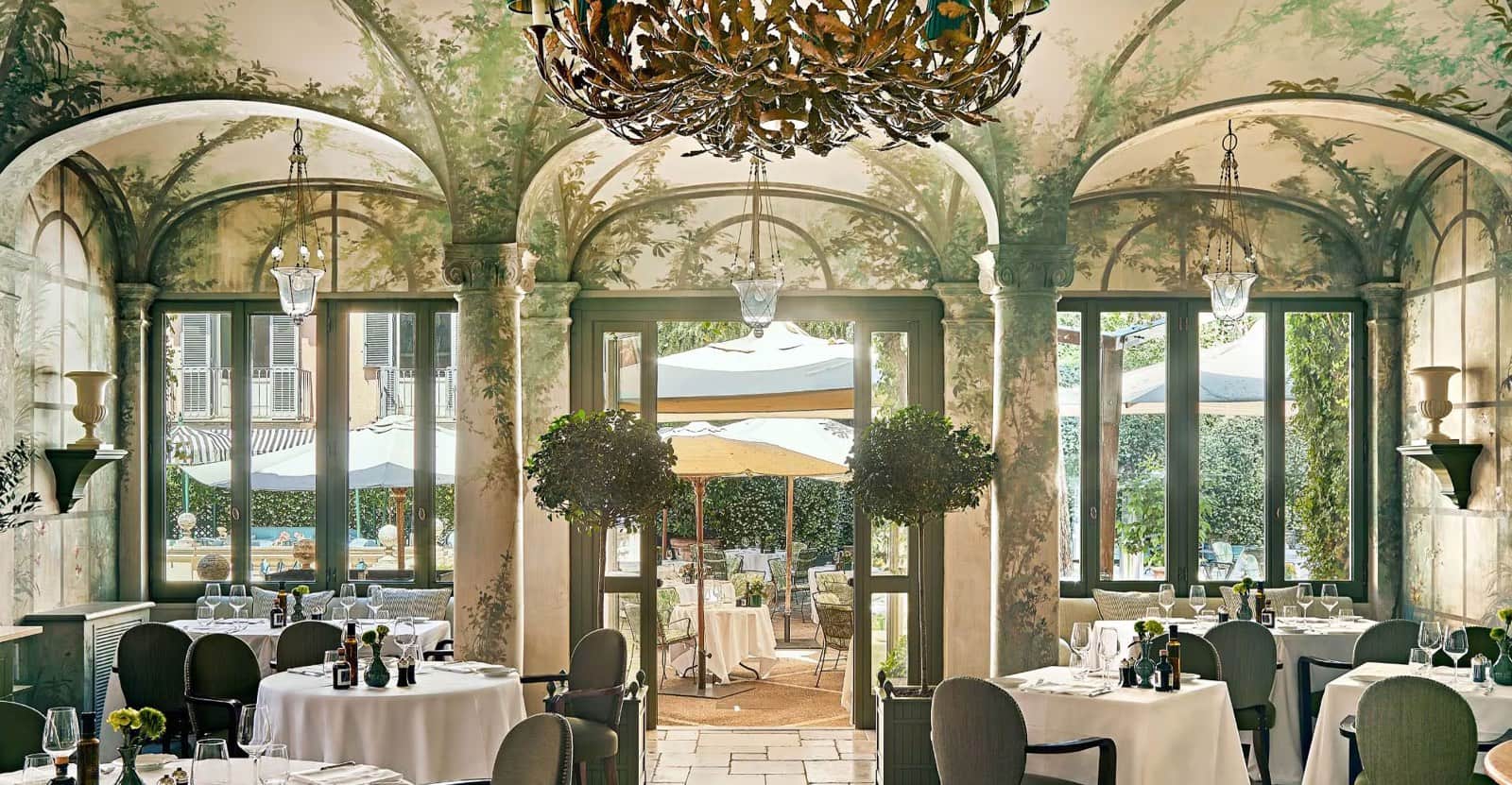 Sala con soffitto a volta affrescato a motivi di foglie, tavoli rotondi apparecchiati e porte a vetri aperte su un giardino con ombrelloni.