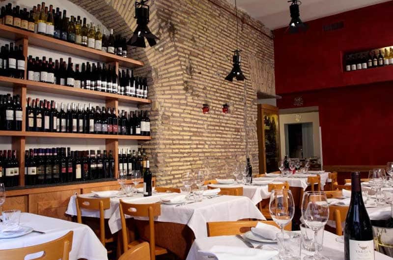 Sala con parete in mattoni ad arco, scaffali di bottiglie di vino e tavoli apparecchiati con tovaglie bianche.