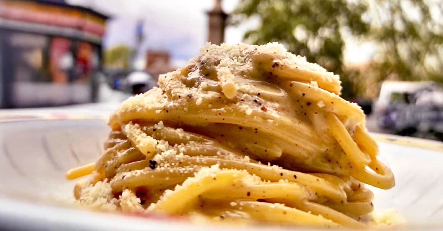 Piatto di spaghetti conditi con crema di formaggio e pepe nero, in primo piano su uno sfondo di strada sfocato.
