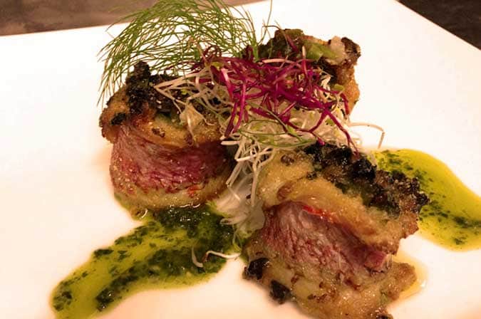 Involtini di carne rosata al centro con crosta scura, su piatto bianco con salsa verde ed erbe fresche viola sopra.