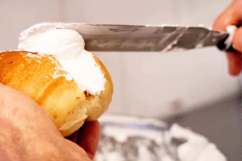 Una mano spalma panna montata su un panino dorato con una spatola metallica.