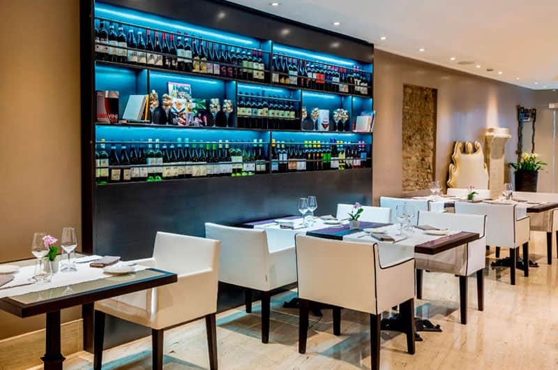 Sala ristorante con parete di bottiglie di vino illuminata e tavoli con poltroncine bianche.