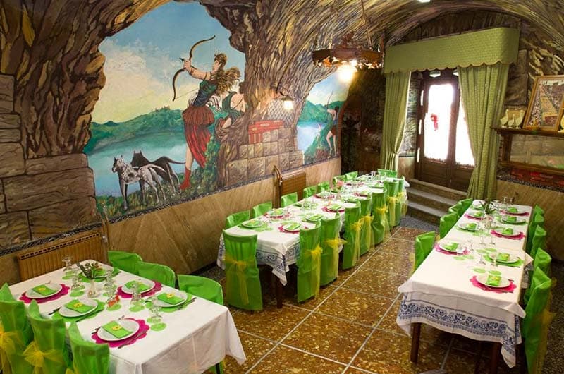 Sala da ristorante con soffitto a volta in pietra, tavoli apparecchiati e murale di una scena di caccia sulla parete.