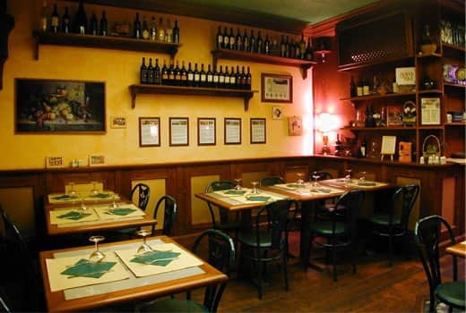Sala da pranzo con pareti gialle, mensole di bottiglie di vino e tavoli con tovagliette verdi e bicchieri.