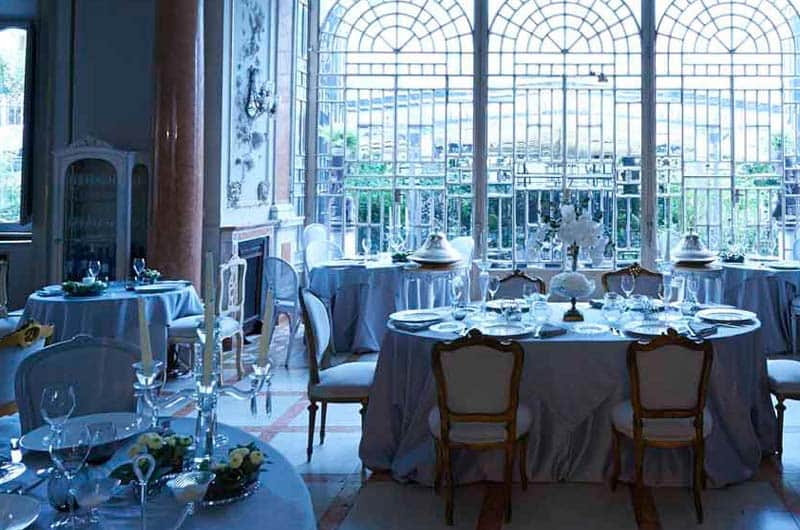 Sala con grandi finestre ad arco, tavoli rotondi apparecchiati con tovaglie grigio-azzurre e sedie in legno chiaro.