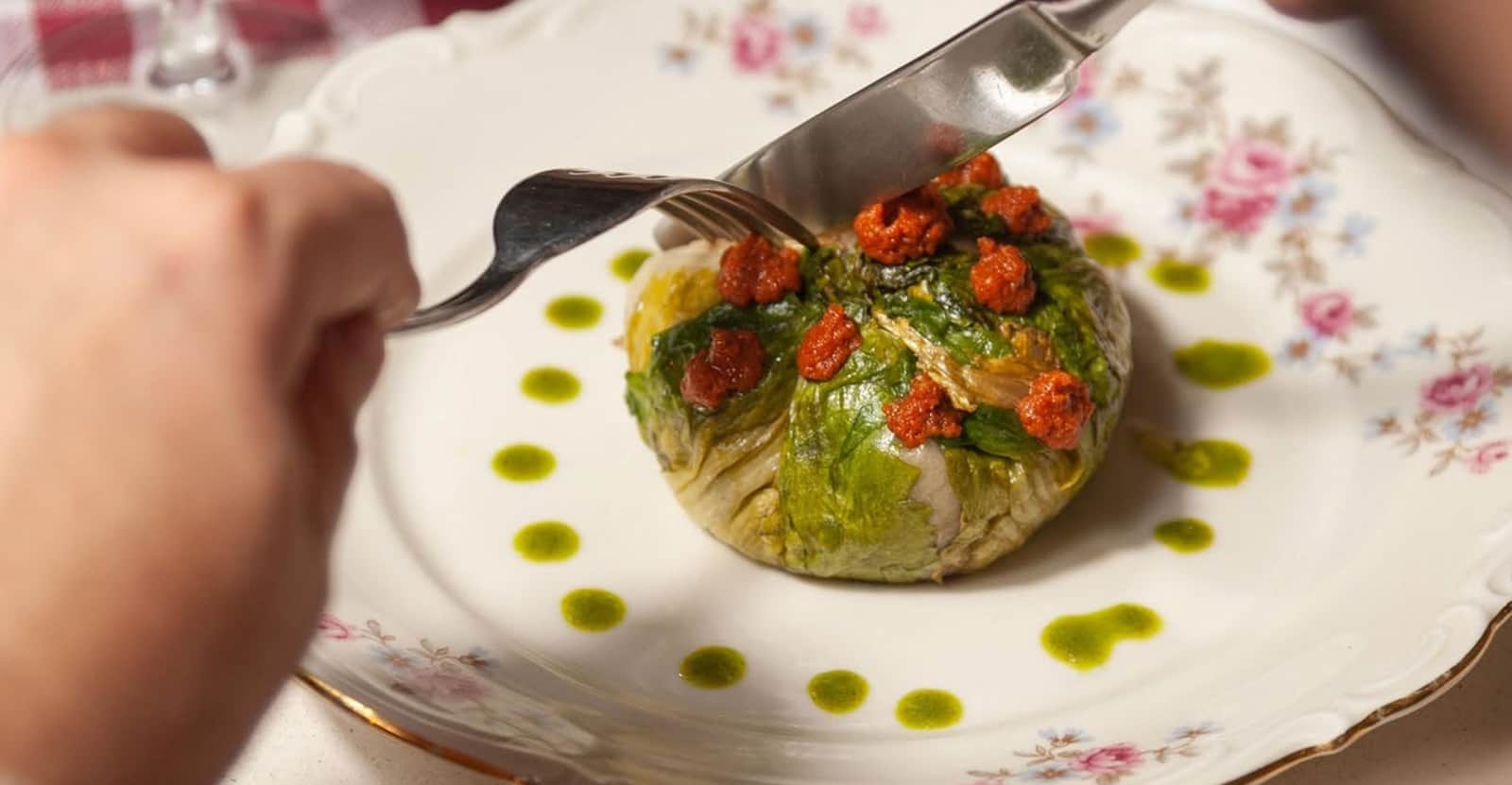Involtino di foglie verdi con piccoli pezzi rossi sopra, tagliato con forchetta e coltello su un piatto decorato.