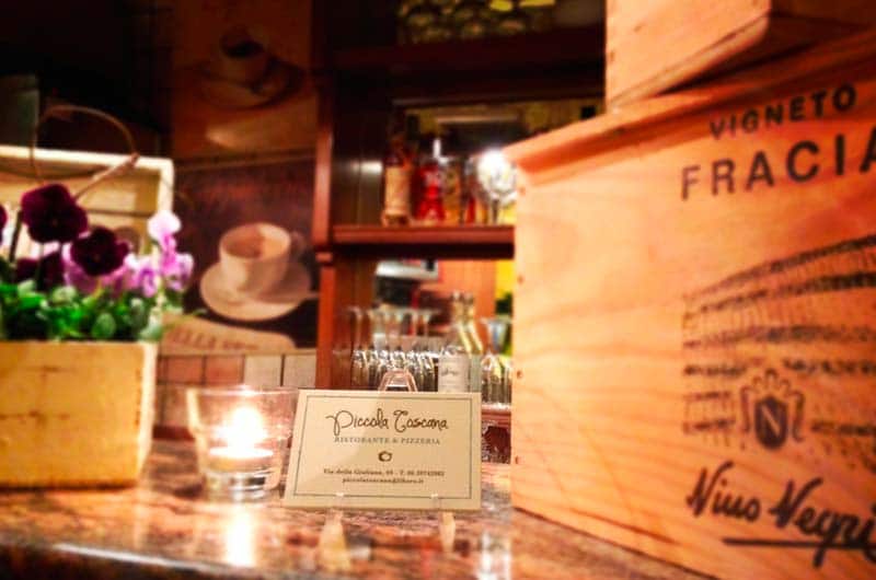 Candela accesa in un bicchiere e cartoncino con scritta 'Piccola Toscana' su un bancone, con una cassa di legno da vino a destra.