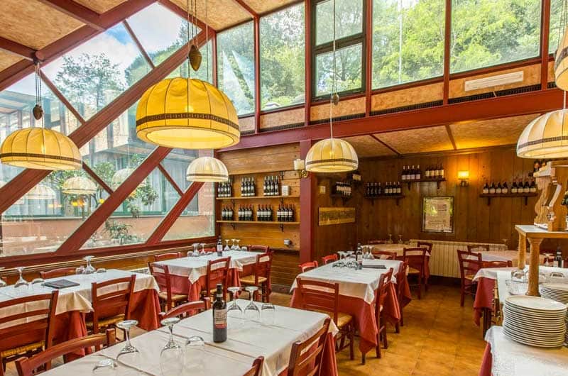Sala con vetrate a capriate rosse, lampadari a campana gialli e tavoli apparecchiati con tovaglie bianche e rosse.