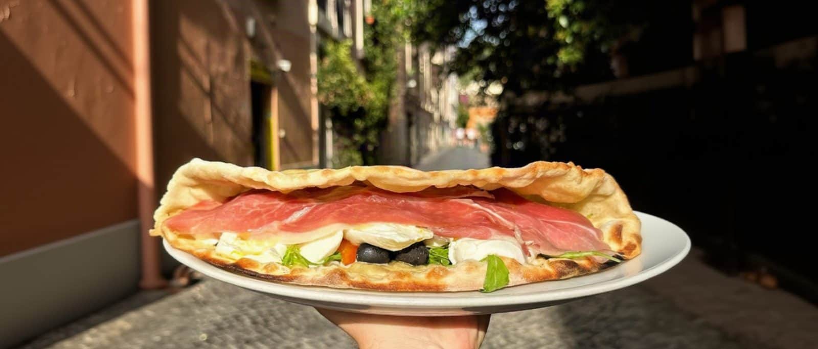 Una mano regge un piatto con una pizza ripiegata a libro, farcita con prosciutto crudo, formaggio bianco e olive nere.