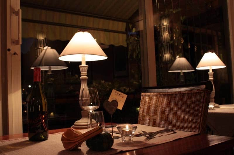 Tavolo apparecchiato di sera con candela accesa, bottiglia di vino e lampade da tavolo illuminate sullo sfondo.