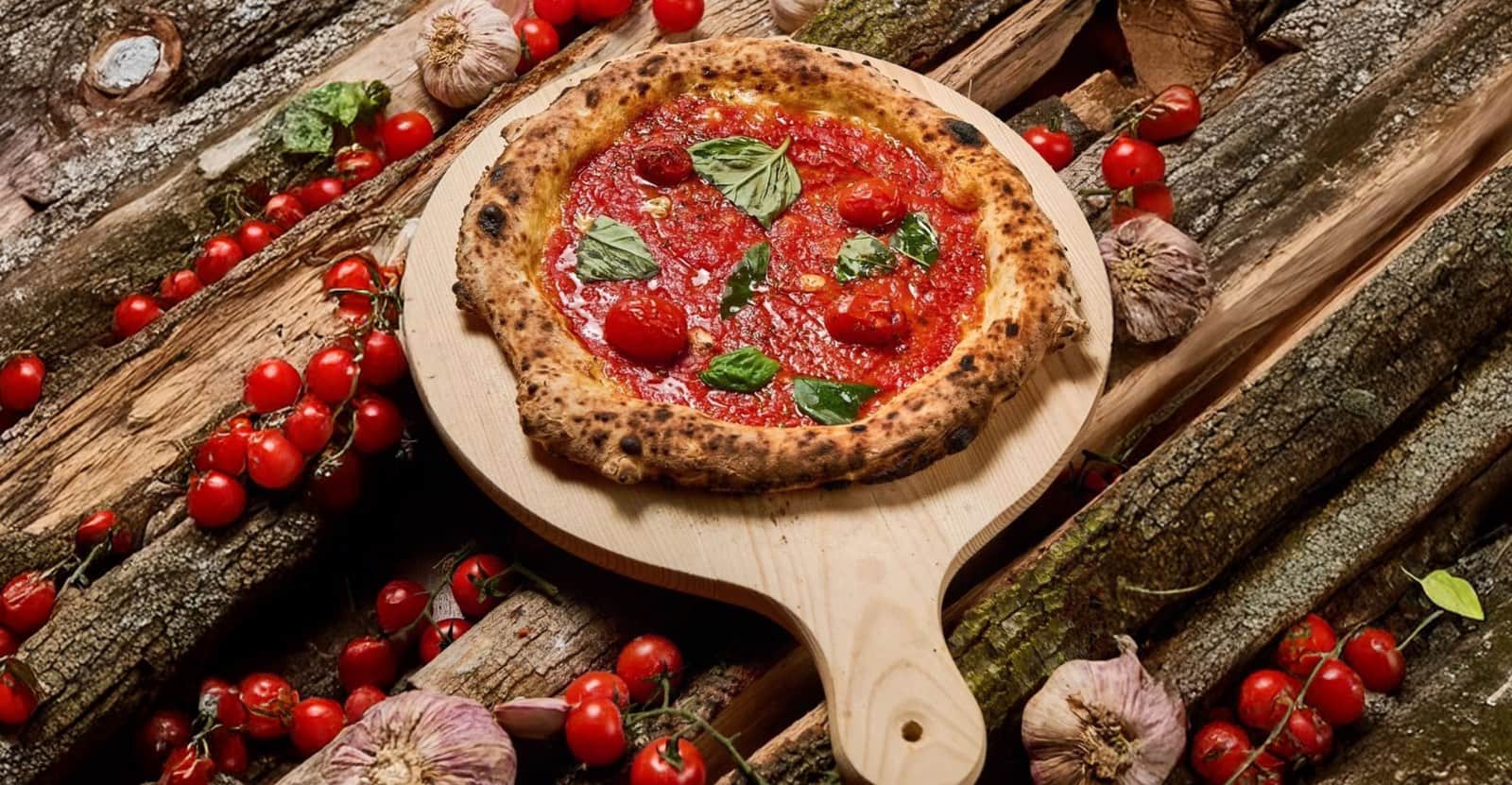 Pizza con bordo alto e bruciacchiato, sugo di pomodoro e basilico, su tagliere di legno tra tronchi e pomodorini.