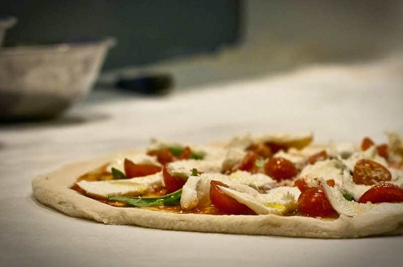 Disco di pasta cruda con pomodorini tagliati, foglie di basilico e pezzi di mozzarella, su un piano chiaro.
