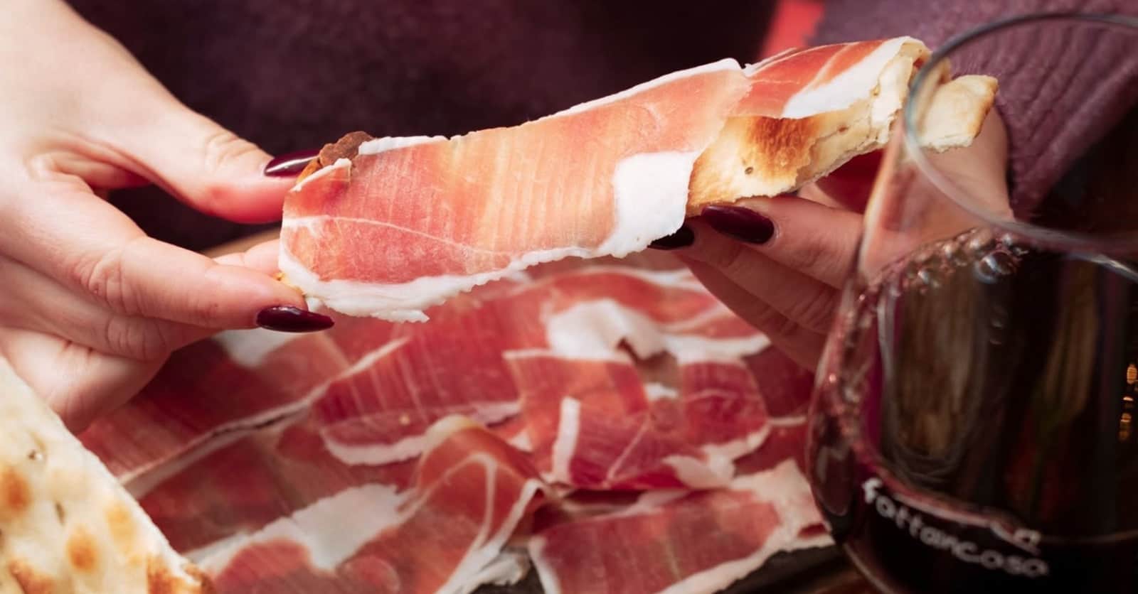 Mani con smalto scuro sollevano una fetta di prosciutto e pane sottile, sopra un vassoio di prosciutto affettato.