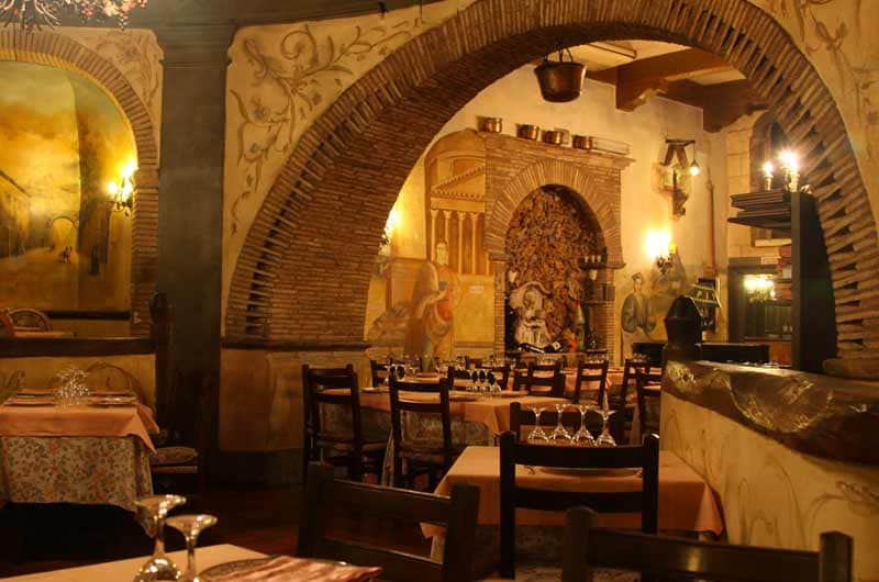 Sala da ristorante con archi in mattoni a vista, tavoli apparecchiati e affreschi murali sullo sfondo.