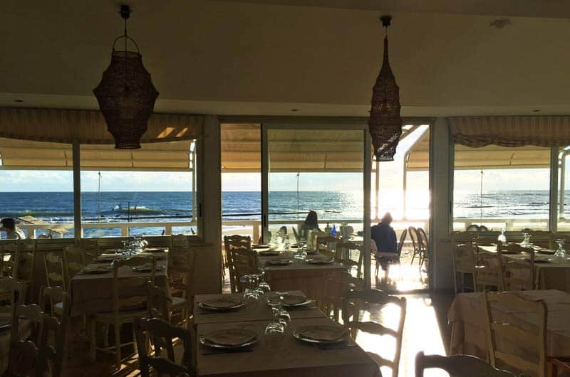 Sala con tavoli apparecchiati e lampade di vimini, davanti a vetrate che si aprono sul mare al tramonto.