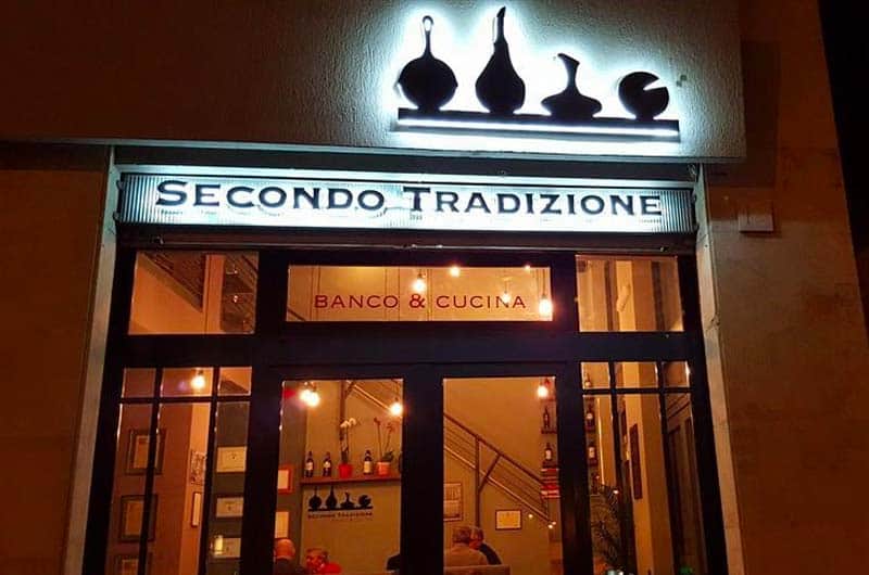 Facciata notturna con insegna luminosa Secondo Tradizione e sagome nere di utensili da cucina sopra l'ingresso.