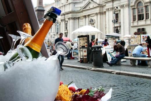 Bottiglia di spumante e calici in un secchiello di ghiaccio, sullo sfondo una piazza con facciata barocca.