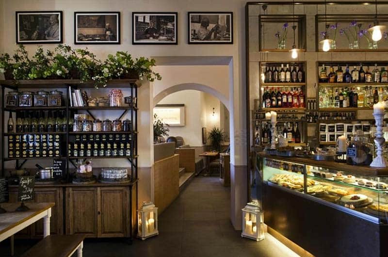 Sala con banco bar in legno scuro e vetrina di dolci, scaffali con bottiglie illuminate e vasi di fiori viola.