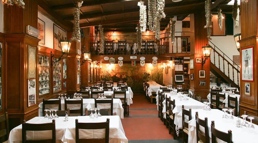 Sala da pranzo in legno con tavoli apparecchiati di bianco, treccine d'aglio appese al soffitto e balconata superiore.