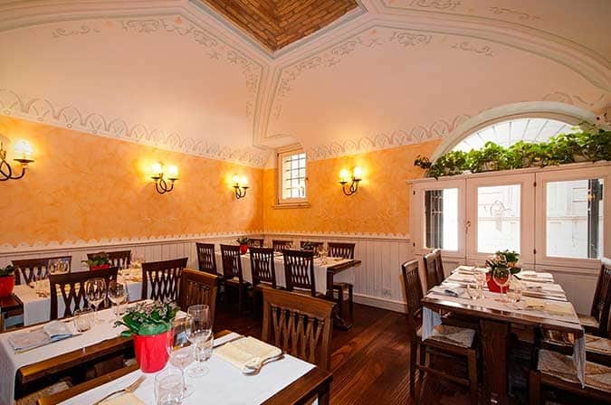 Sala con soffitto a volta affrescato, pareti color ocra, applique in ferro battuto e tavoli apparecchiati con tovaglie bianche.
