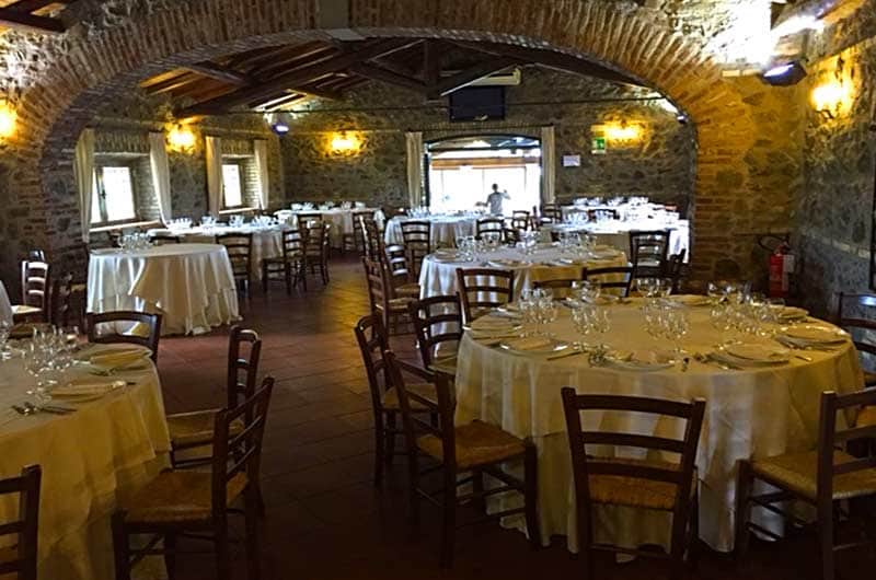 Sala da ristorante con volta in mattoni, tavoli rotondi apparecchiati e sedie in legno con seduta in paglia.