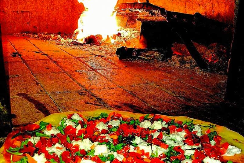 Pizza con pomodori, mozzarella ed erbe verdi in primo piano, sullo sfondo un forno a legna con le fiamme accese.