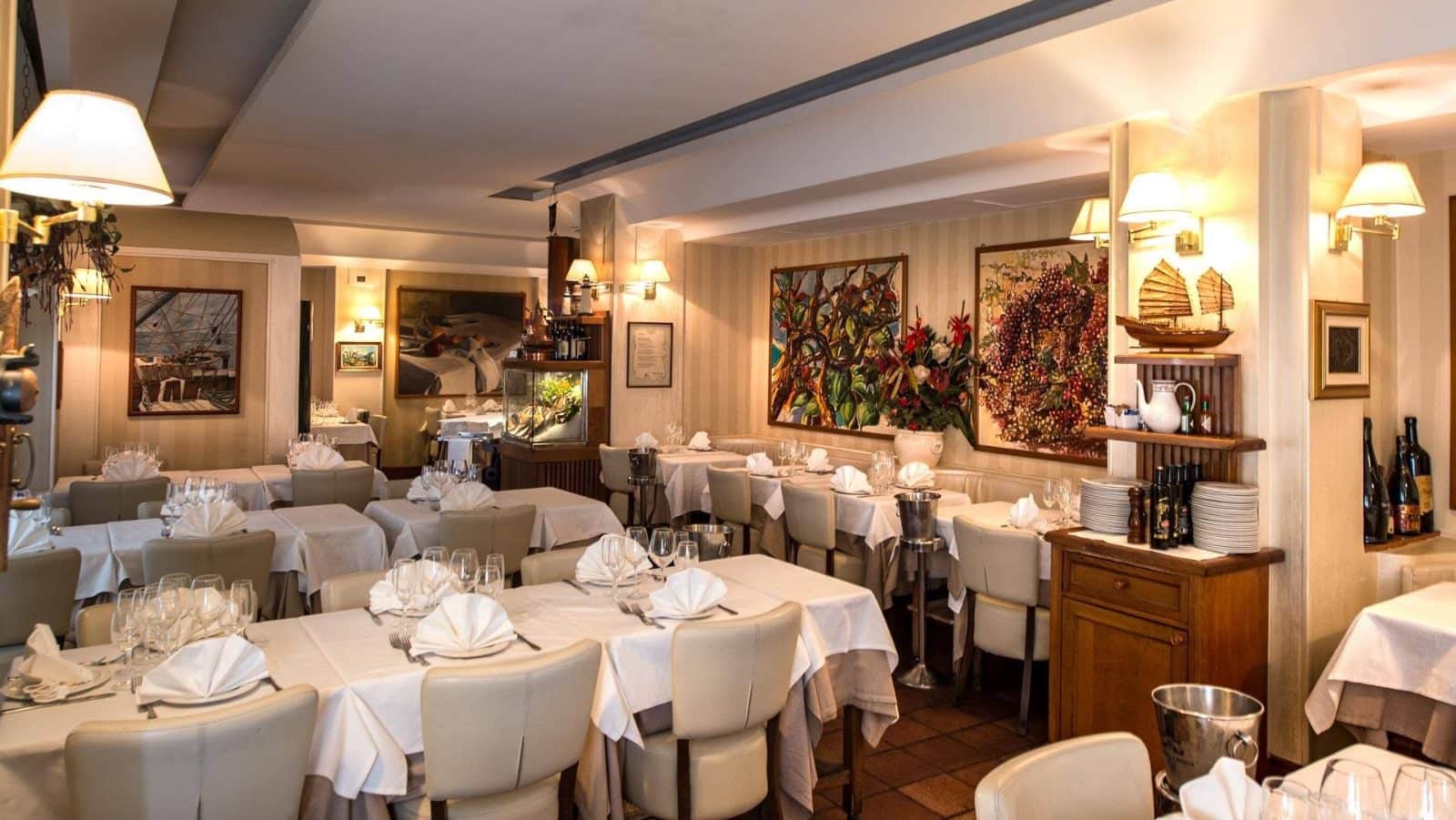 Sala da pranzo con tavoli rotondi apparecchiati in bianco, quadri colorati alle pareti e applique dorate.