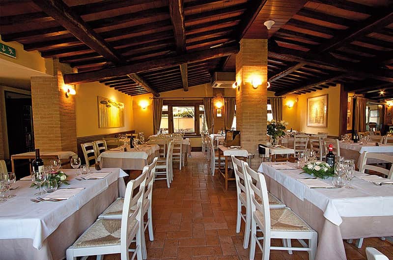 Sala da pranzo con travi di legno a vista, colonne in mattoni e tavoli apparecchiati con tovaglie bianche.