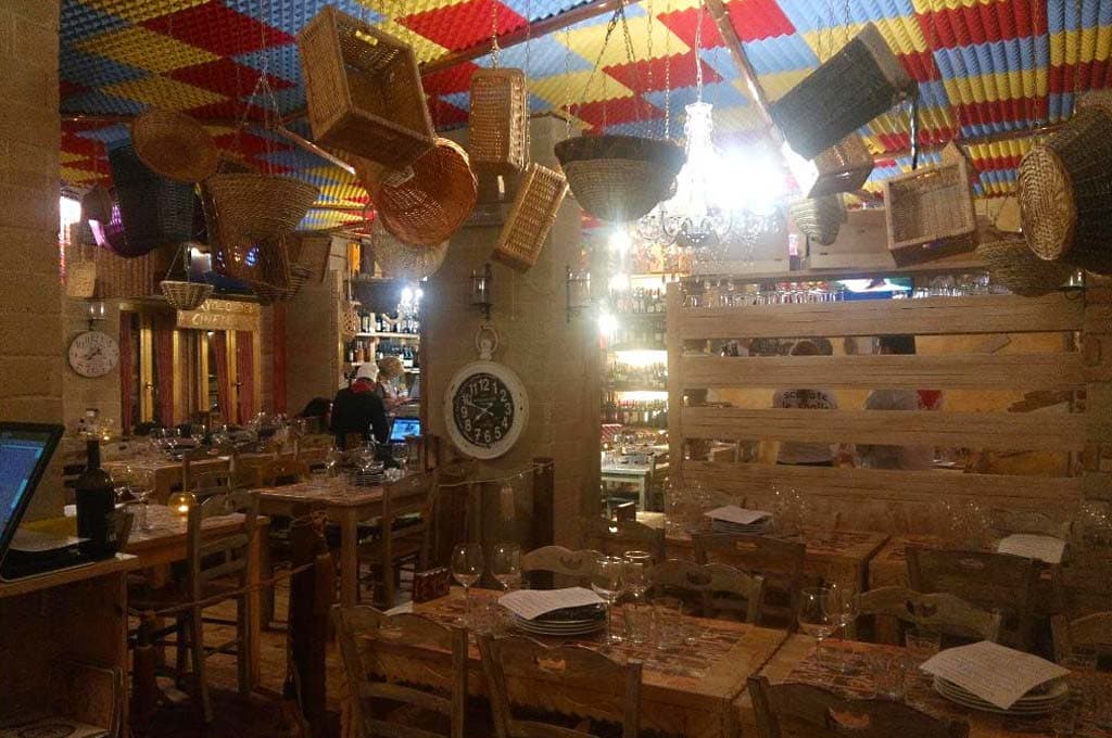 Sala con soffitto a pannelli colorati, ceste di vimini appese e tavoli in legno apparecchiati con bicchieri da vino.