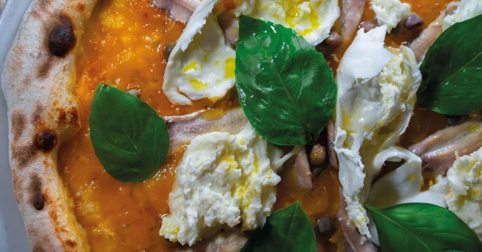 Base di pizza con salsa arancione, mozzarella a lembi, foglie di basilico e filetti di acciuga.