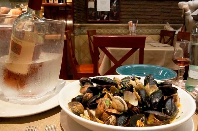 Cozze e vongole in guazzetto rosso in una ciotola bianca, con un secchiello del vino e un calice sullo sfondo.