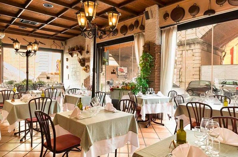 Sala con tavoli apparecchiati, sedie in legno scuro, pareti in mattoni e grandi finestre su strada.