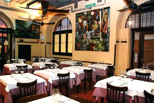 Sala da ristorante con tavoli apparecchiati in bianco e bordeaux, sedie scure e un grande dipinto colorato sulla parete.
