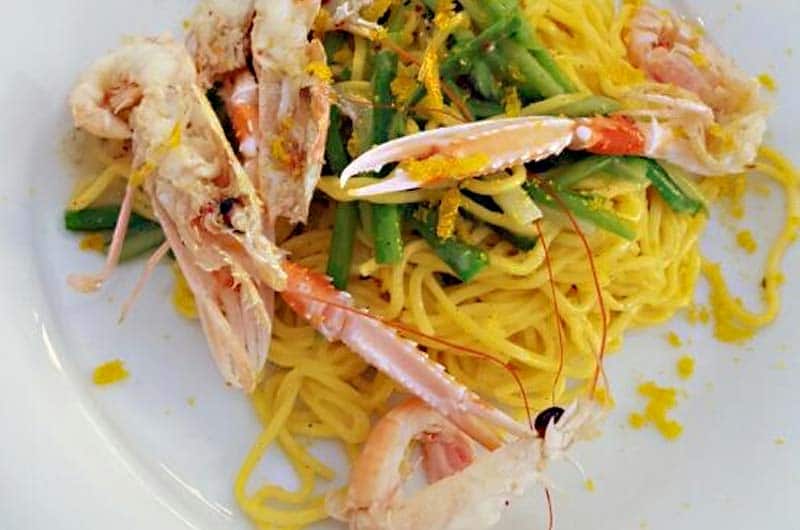 Spaghetti gialli con code di gambero, chele di crostaceo e fagiolini verdi su piatto bianco.