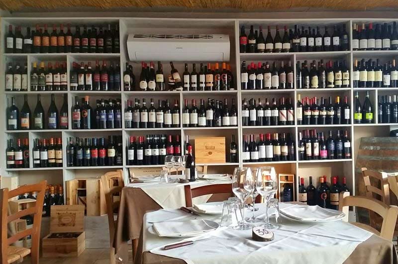 Scaffali pieni di bottiglie di vino coprono la parete, con un tavolo apparecchiato con calici in primo piano.