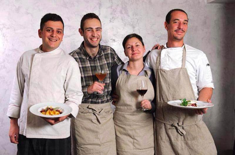 Quattro persone in tenuta da cucina sorridono affiancate, due con piatti in mano e due con bicchieri di vino.