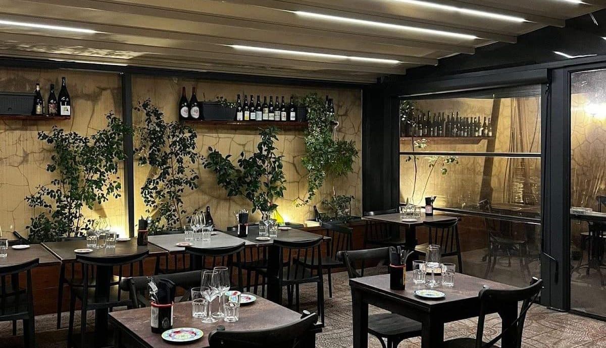 Sala con tavoli in legno scuro apparecchiati, piante rampicanti su parete chiara e bottiglie su mensole.