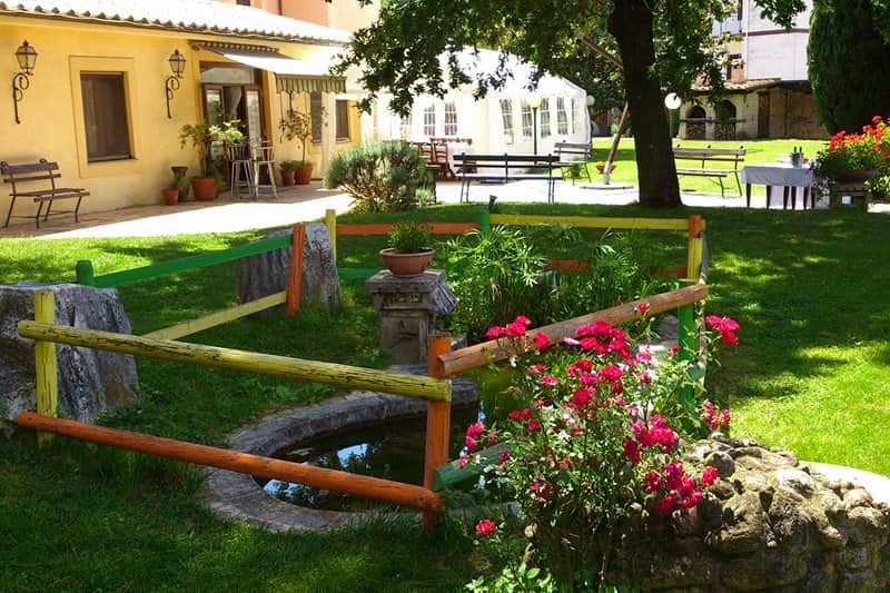 Giardino con staccionata colorata attorno a una fontana in pietra e cespugli di rose rosa e rosse.