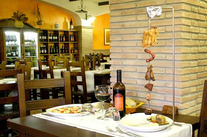 Spiedo verticale con pezzi di carne grigliata accanto a un tavolo con pizza, vino e sala del ristorante sullo sfondo.