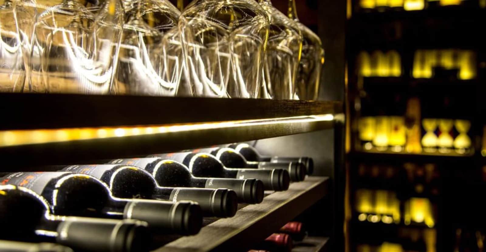 Bottiglie di vino sdraiate su una rastrelliera in legno, con calici appesi capovolti sopra.