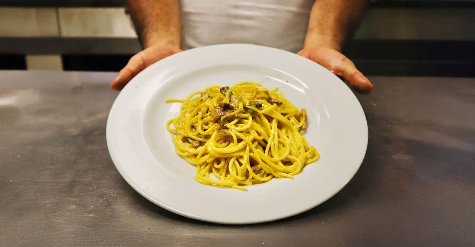 Piatto di spaghetti conditi con sugo giallo e pezzetti scuri, porto in avanti da un cameriere.