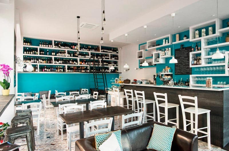 Sala di un locale con pareti turchesi coperte di scaffali di bottiglie di vino e bancone bar in legno scuro.