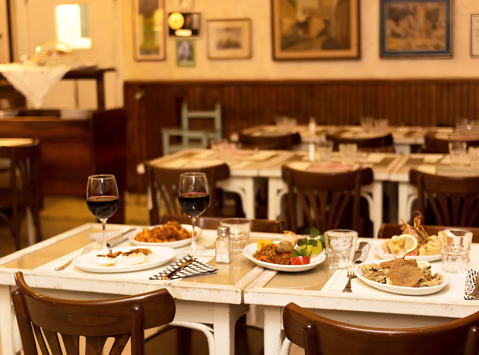 Tavolo apparecchiato con piatti di carne e verdure, due bicchieri di vino rosso, sala con tavoli in legno sullo sfondo.