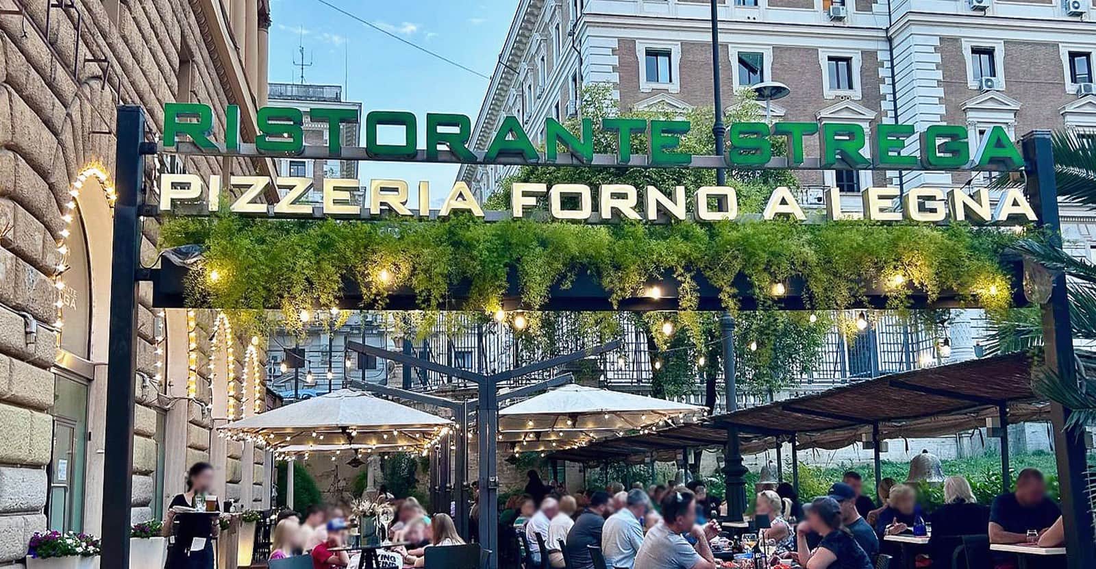Insegna verde e bianca con piante pensili sopra tavoli all'aperto con ombrelloni bianchi e persone sedute.