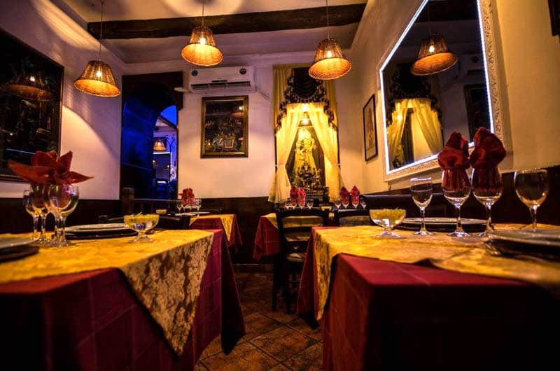 Sala da ristorante con tavoli imbanditi in rosso e oro, tovaglioli decorativi rossi e lampade a cesto sospese dal soffitto.