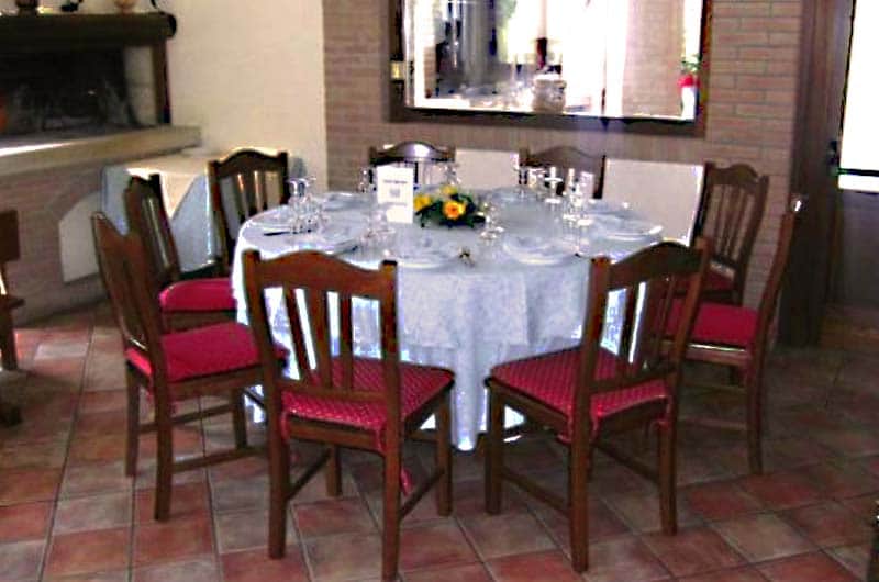 Tavolo rotondo apparecchiato con tovaglia bianca, composizione di fiori gialli al centro e sedie in legno con seduta rossa intorno.